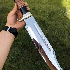 May include: A large, polished stainless steel knife with a curved blade. The handle features a black and brown leather grip, accented with gold-colored metal. The knife is held against a backdrop of green grass.