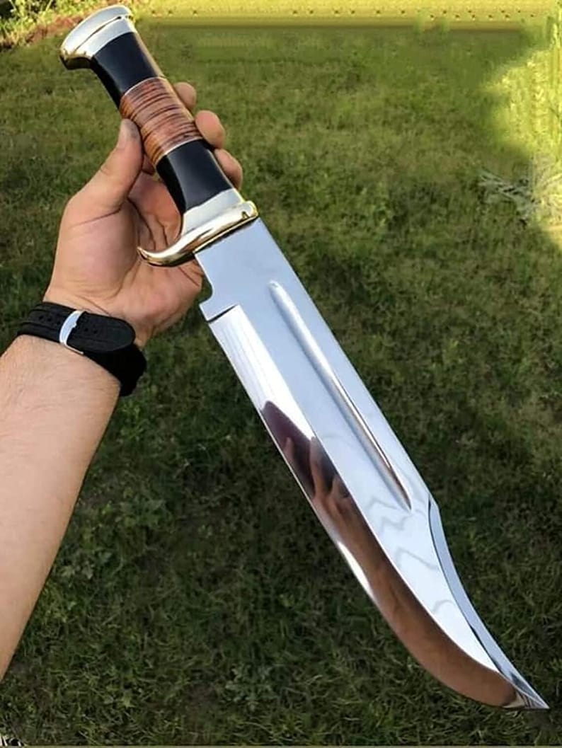 May include: A large, polished stainless steel knife with a curved blade. The handle features a black and brown leather grip, accented with gold-colored metal. The knife is held against a backdrop of green grass.