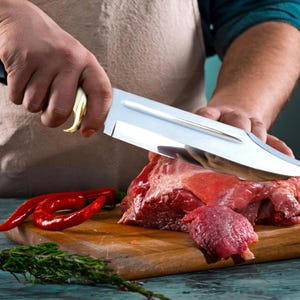 May include: A large, stainless steel butcher knife with a gold-colored handle is cutting through a piece of raw red meat on a wooden cutting board. Red chili peppers and green herbs are also on the board.