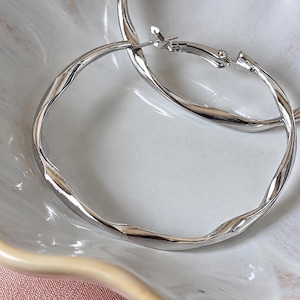 May include: A pair of silver-toned hoop earrings featuring a twisted, textured metal design. The earrings have a polished, reflective finish and include a hinged post closure mechanism that secures the circular frame in place.