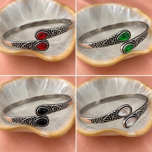 May include: A collection of four open-ended cuff bracelets made of antiqued silver-tone metal. Each bracelet features two teardrop-shaped stones at the ends, available in red, green, black, or white, with intricate embossed scrollwork and dot detailing.