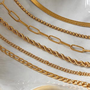 May include: A collection of five gold-toned metal chains featuring various link styles, including a flat herringbone chain, a round bead chain, an elongated oval link chain, a twisted rope chain, and a classic curb chain, all arranged in parallel.