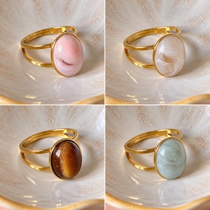 Op de afbeelding: Een collectie van vier goudkleurige ringen, elk voorzien van een gladde, ovaalvormige cabochonsteen. De stenen vertonen duidelijke gemarmerde of chatoyante patronen in tinten van zachtroze, crèmekleurig beige, diep amberbruin en licht muntgroen.