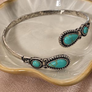 May include: An open-ended silver-toned cuff bracelet featuring a weathered metal finish. The two ends are decorated with ornate metal detailing and inlaid with four teardrop-shaped turquoise stones that display natural brown veining throughout.