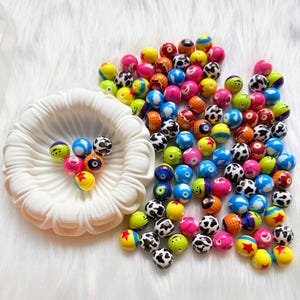 Könnte beinhalten: Eine Sammlung runder, mehrfarbiger Perlen mit verschiedenen Mustern, darunter Kuhflecken, einfarbige Designs und Sternmotive, wobei eine kleine Gruppe dieser Perlen in einem weißen, gewellten, schalenförmigen Behälter liegt.