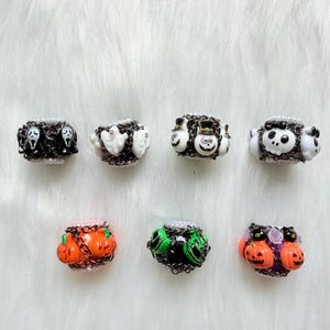 May include: Seven decorative rings featuring Halloween-themed charms including ghost faces, white ghosts, top-hatted skulls, white skulls, orange pumpkins, green monsters, and black cats with orange pumpkins, all accented with small white beads and chains.