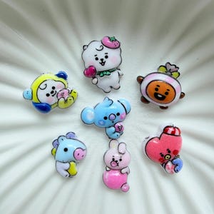 Pode incluir: Uma coleção de sete pequenos berloques brilhantes e coloridos com vários personagens de desenhos animados, incluindo uma criatura branca com um chapéu de morango, um coala azul, um personagem em forma de coração rosa e outros designs estilizados.