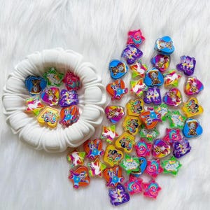 May include: A collection of small, colorful plastic charms in various shapes including hearts and stars. The charms feature vibrant, multi-colored graphic prints of cartoon animals and rainbows. Some charms are inside a white, fluted bowl.