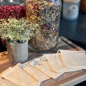 May include: Six natural muslin tea bags with drawstring closures, arranged on a wooden surface. Two glass jars filled with dried herbs and flowers are in the background.