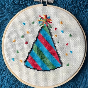May include: A white fabric with a colorful party hat embroidered on it. The hat is blue, red, and green with a yellow and orange star on top. The fabric is in a wooden embroidery hoop.
