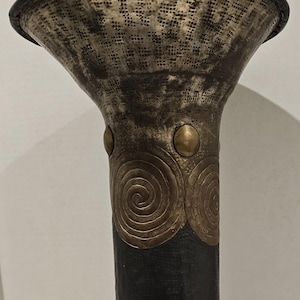 May include: A tall, decorative vase with a flared top and a textured, metallic finish. The vase features a dark, cylindrical body adorned with brass accents, including circular designs and small studs. The base has three brass feet.