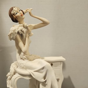 May include: A cream-colored figurine of a woman in a flowing gown, seated on a decorative chair. The figurine has a feathered shawl and a small nightstand. The base is dark brown, with the text "Giuseppe Armani".