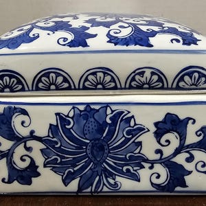 May include: A rectangular, lidded ceramic box with a white base and blue floral designs. The lid features a repeating pattern of stylized flowers, while the body showcases a large floral motif with swirling leaves. The box is a decorative item.