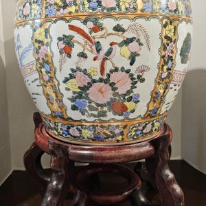 May include: A large, ornate ceramic planter with a detailed floral and bird design, set on a dark wooden stand. The planter features a white base with colorful flowers, birds, and fruit motifs, accented by gold trim.
