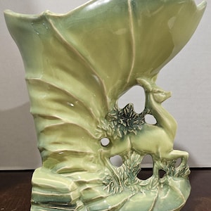 May include: A light green ceramic vase featuring a wide, flared opening and a sculpted deer figure standing amidst foliage on the base. The piece has a glossy finish with darker green shading in the recessed areas of the relief design.