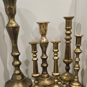 May include: Set of seven antique brass candle holders of varying heights and styles. The candle holders are all made of brass and have a polished finish.