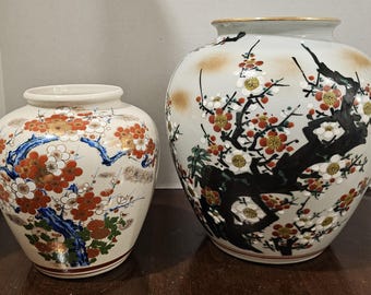May include: Two ceramic vases with floral designs. The smaller vase has a cream background with red, white, and gold flowers and blue branches. The larger vase has a light green background with a black tree and red, white, and gold flowers.