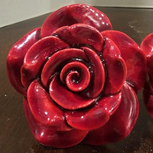 May include: Close-up of a glossy, deep red rose. The petals are intricately detailed, with a dark, almost black center. The rose appears to be made of a hard material, possibly ceramic or resin, and has a high-shine finish. The overall effect is elegant and decorative.