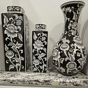 May include: Three black and white ceramic vases with floral designs. The tallest vase is a pear-shaped vase with a wide opening. The two shorter vases are square and have a lid.