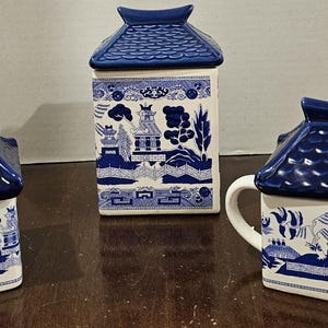 May include: A three-piece ceramic tea set featuring a blue and white Willow pattern. The set includes a teapot, a sugar bowl, and a creamer, each with a blue roof-shaped lid. The teapot has a handle and spout.