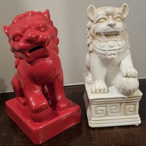 May include: Two ceramic Foo Dog statues. One is a glossy red, the other is off-white. The red Foo Dog is seated on a rectangular base. The off-white Foo Dog is seated on a square base and holds a ball.