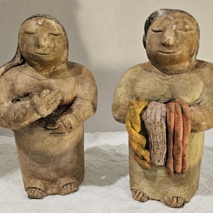 May include: Two ceramic figurines of figures with closed eyes. One figure has hands clasped in front, the other holds a bundle of fabric in shades of yellow, tan, and orange. Both figures are in neutral tones.