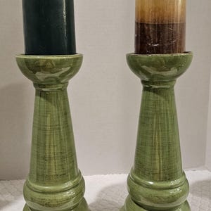 May include: Two decorative candle holders with candles. The holders are green with a distressed finish. One candle is dark green, and the other is brown and orange. The candle holders are on a white surface.