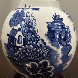 May include: A white ceramic vase with a blue and white landscape design. The design features pagodas, trees, and foliage. The vase has a rounded shape with a wide opening.