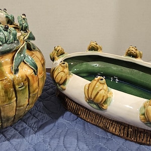 May include: Two ceramic planters. One is round, resembling bamboo, with green and yellow tones, and green frog figurines. The other is an oval planter with a white exterior, green interior, and frog figurines around the rim.