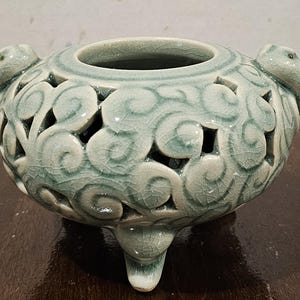 May include: A celadon-colored ceramic incense burner with a round body and three short legs. The body features intricate cut-out designs and two handles. The surface has a crackle glaze.