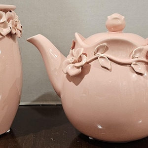 May include: A pink ceramic teapot and vase set. The teapot has a curved handle, a spout, and a lid with a rose-shaped knob. Both pieces are decorated with floral accents.