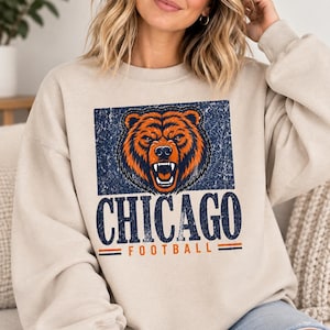 May include: A beige crewneck sweatshirt featuring a distressed graphic of a snarling bear head in orange and dark blue. Below the bear, the text reads CHICAGO in large dark blue letters, with the word FOOTBALL in orange underneath.