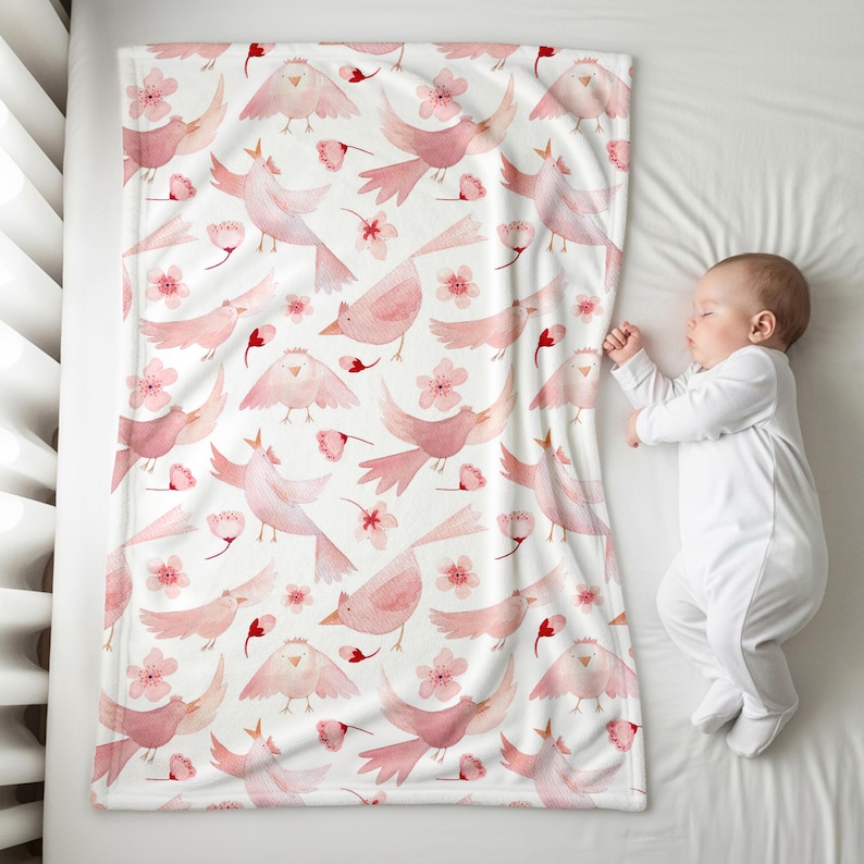 K&ouml;nnte beinhalten: Eine wei&szlig;e Babydecke mit einem sich wiederholenden Muster aus rosa V&ouml;geln und Blumen. Die Decke ist rechteckig und bedeckt ein Baby in einem wei&szlig;en Strampler. Die V&ouml;gel sind in verschiedenen Posen, und die Blumen bl&uuml;hen.