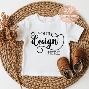 Pode incluir: Camiseta branca para crianças pequenas com o texto "YOUR design HERE" em escrita preta. A camisa é exibida em um tapete tecido com sapatos de bebê de couro marrom. Um raminho de flores secas está à esquerda.