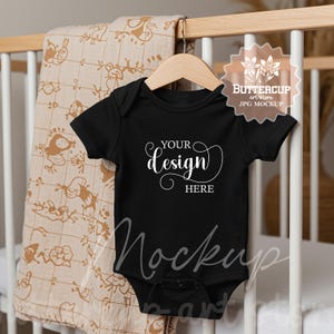 Könnte beinhalten: Schwarzer Baby-Body-Mockup mit dem Text "YOUR design HERE" auf weißem Hintergrund. Der Body hängt an einem Holzbügel vor einem weißen Babybett.
