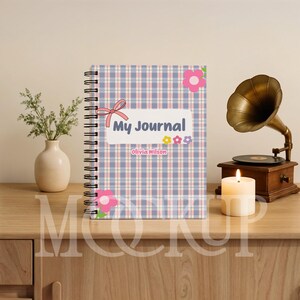 May include: A spiral-bound journal with a plaid pattern in blue, pink, and white. The cover features the text "My Journal" and the name "Olivia Wilson", along with floral and bow accents. A vintage gramophone and lit candle are in the background.