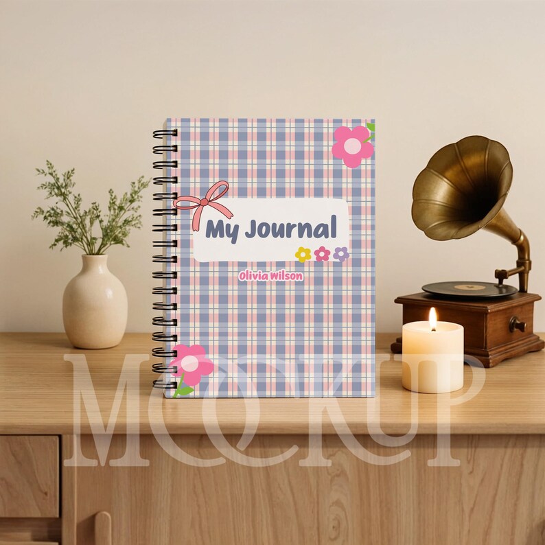 May include: A spiral-bound journal with a plaid pattern in blue, pink, and white. The cover features the text "My Journal" and the name "Olivia Wilson", along with floral and bow accents. A vintage gramophone and lit candle are in the background.
