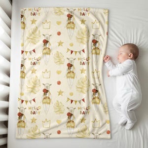 K&ouml;nnte beinhalten: Eine cremefarbene Babydecke mit einem sich wiederholenden Muster eines Lamms mit Latzhose, Kronen, Sternen und dem Text "HELLO BABY". Die Decke hat auch Bl&auml;tter, Herzen und Wimpel im Aquarellstil. Ein schlafendes Baby liegt neben der Decke.