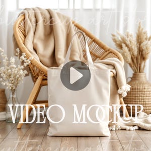 Puede incluir: Una bolsa de lona blanca con un icono de botón de reproducción en la parte delantera. La bolsa está sentada en un suelo de madera delante de una silla de mimbre con una manta de lana beige.