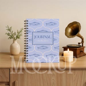 May include: A spiral-bound journal with a light blue cover featuring a repeating fish pattern. The journal has the word "JOURNAL" and the name "Claudia Alves" printed on the front. A lit candle and a vintage gramophone are in the background.