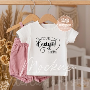 May include: White short-sleeved t-shirt mockup with the text "YOUR design HERE" on a hanger. The t-shirt is paired with pink shorts and is hanging in a nursery setting.