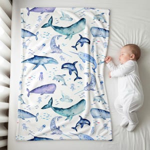 K&ouml;nnte beinhalten: Eine wei&szlig;e Babydecke mit einem Aquarelldesign von Walen, Delfinen, Narwalen und Quallen in Blau- und Lilat&ouml;nen. Die Decke liegt in einem Kinderbett neben einem schlafenden Baby.