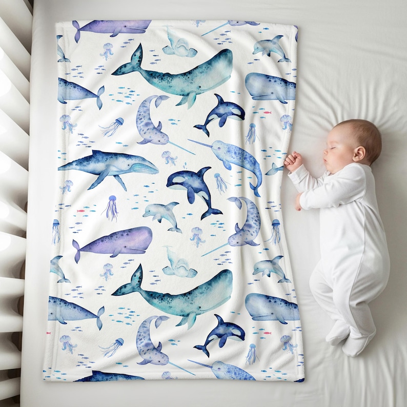 K&ouml;nnte beinhalten: Eine wei&szlig;e Babydecke mit einem Aquarelldesign von Walen, Delfinen, Narwalen und Quallen in Blau- und Lilat&ouml;nen. Die Decke liegt in einem Kinderbett neben einem schlafenden Baby.