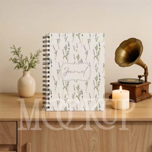 May include: A spiral-bound journal with a lavender floral pattern and the word "Journal" in script. A vintage gramophone, a lit candle, and a small vase with greenery are on a wooden surface.