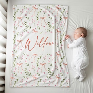 K&ouml;nnte beinhalten: Eine wei&szlig;e Babydecke mit einem floralen Muster aus rosa Schleifen, Blumen und gr&uuml;nen Bl&auml;ttern. Der Name "Willow" ist in korallfarbener Schrift geschrieben. Die Decke liegt auf einer wei&szlig;en Oberfl&auml;che neben einem schlafenden Baby.