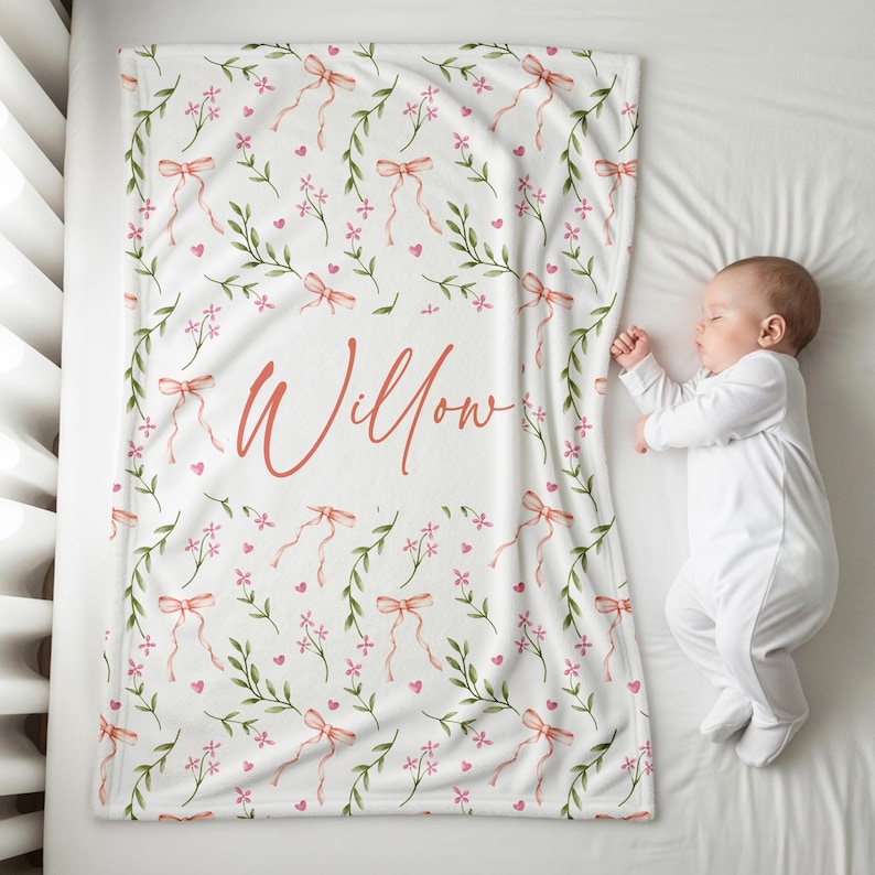 K&ouml;nnte beinhalten: Eine wei&szlig;e Babydecke mit einem floralen Muster aus rosa Schleifen, Blumen und gr&uuml;nen Bl&auml;ttern. Der Name "Willow" ist in korallfarbener Schrift geschrieben. Die Decke liegt auf einer wei&szlig;en Oberfl&auml;che neben einem schlafenden Baby.