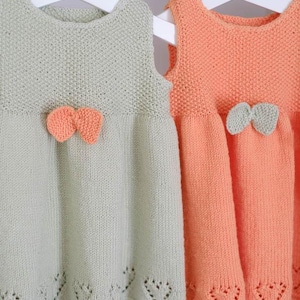 Può includere: Due vestitini per neonati lavorati a maglia su grucce bianche. Uno è verde chiaro con un fiocco arancione, l'altro è color pesca con un fiocco verde chiaro. Entrambi gli abiti hanno un motivo a forma di cuore sull'orlo.