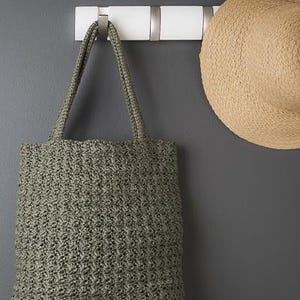 May include: A sage green woven tote bag hangs on a white wall hook next to a straw sun hat. The bag has a rectangular shape with two handles. The hat is round with a wide brim. The wall is painted a dark gray.