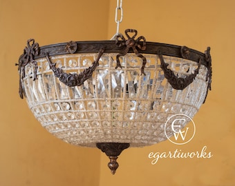 May include: An antique chandelier with a bronze-colored metal frame and clear crystal accents. The chandelier has a bowl shape with decorative bows and floral details. The crystals are arranged in a layered pattern, creating a sparkling effect. The fixture is suspended from a chain.