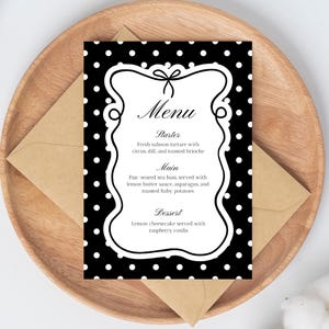 Peut inclure: Une carte de menu en noir et blanc avec un fond à pois et un cadre blanc décoratif. Le menu énumère une entrée, un plat principal et un dessert. La carte est posée sur une assiette en bois avec une enveloppe brune.